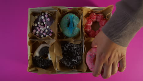 a hand with a gray sleeve places a pink frosted donut with rainbow sprinkles into the final slot of a six-count delivery box, completing a colorful assortment against a bright pink background.