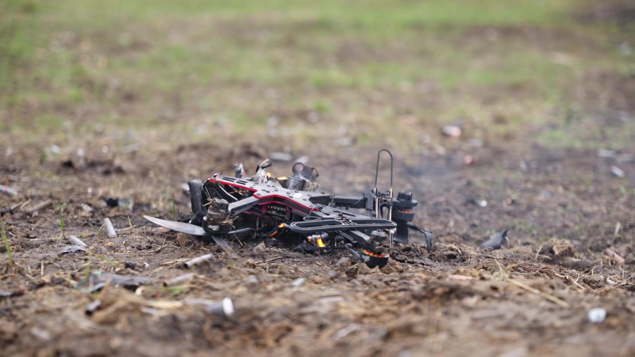 Drone FPV carbonizzato giacente su terreno sterrato con leggero fumo in un campo aperto.