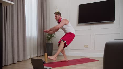 Side-view-of-a-confident-man-with-brown-hair-with-an-athletic-build-and-stubble-wearing-red-wireless-headphones-in-a-white-T-shirt-and-red-shorts-doing-squats-on-one-leg-on-the-carpet-during-his-morning-exercises-at-home-in-a-modern-apartment-in-the-morning