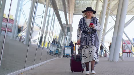 Abuela-Turista-Senior-Caminando-Por-El-Pasillo-Del-Aeropuerto-Internacional,-Usando-Teléfono-Móvil,-Enviando-Mensajes-De-Texto