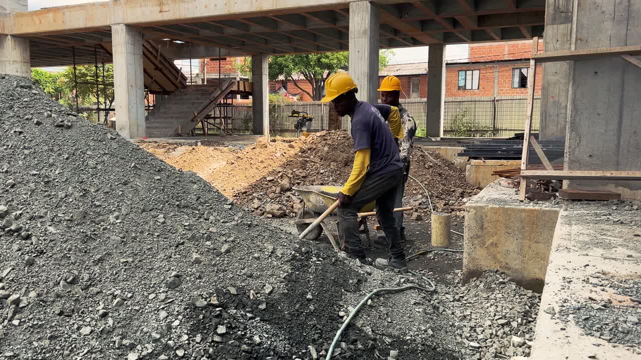 Two construction workers labor at a building site, with one using a shovel to move rocks as part of the construction process.