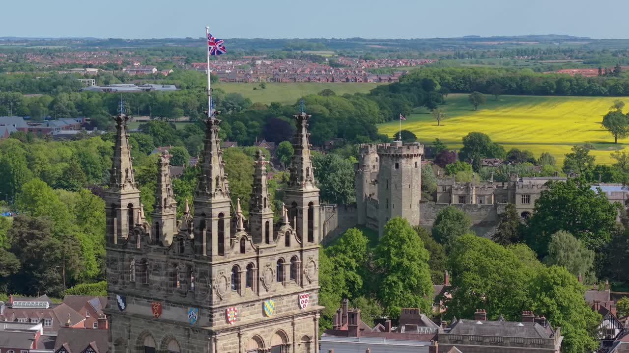 Drohnen-Orbit um den Glockenturm der St. Mary’s Church in Warwick mit wehender Englandflagge, der allmählich das Verlies von Warwick Castle und die umliegende historische Stadtlandschaft enthüllt.