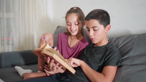 interactive learning session between friends as they sit on couch reading book together with joy, boy points at page while girl smiles holding pen, smartphone placed behind them on sofa armrest