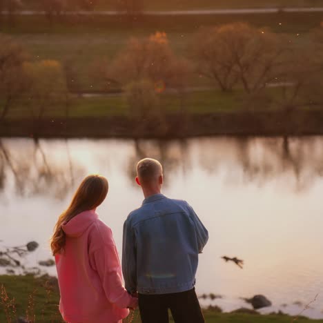 young man and woman looking at the setting sun. romantic couple holding hands while standing on the hill over the river at sunset. rear view