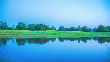 shot of lake surrounded by green vegetation with camp fire going on in distance from night to morning in timelapse