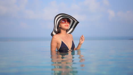 Bonita-Mujer-Asiática-Con-Sombrero-Y-Gafas-De-Sol-Dentro-De-La-Piscina-Infinita-Girando-Lentamente-Hacia-La-Cámara,-Concepto-De-Turismo-En-Cámara-Lenta