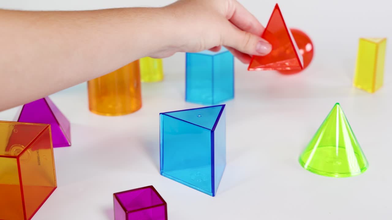 Hands interact with vibrant 3D geometric shapes on a white surface, demonstrating playful learning and spatial arrangement