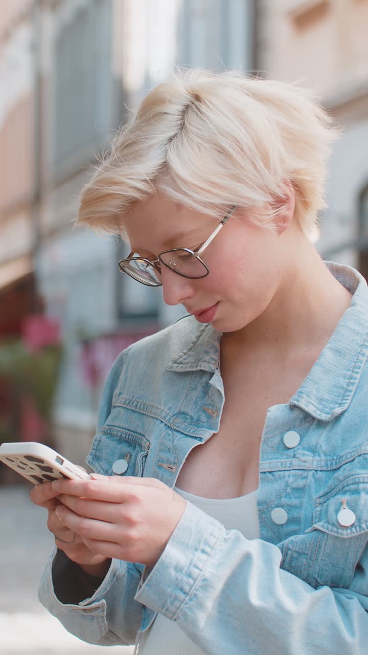 Smiling blond woman using smartphone typing texting scrolling social media app on city town street