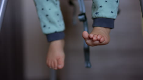 bare little feet of a little child wearing blue pants. baby sitting in a feeding chair and waving legs cheerfully. close up. blurred backdrop.