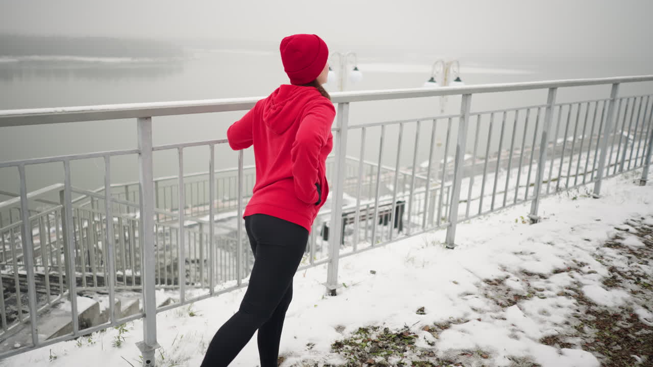 겨울 동안 야외에서 운동하는 여성의 측면 뷰, 균형을 잡기 위해 철 레일링을 들고, 눈 인 땅, 안개 분위기, 멀리 있는 강, 배경에 있는 조명 기둥으로 둘러싸여 있습니다.