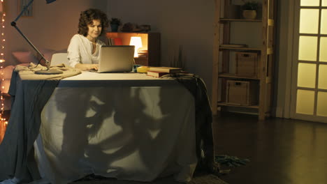 smiling young woman working from home with laptop computer and looking after two little children playing under desk