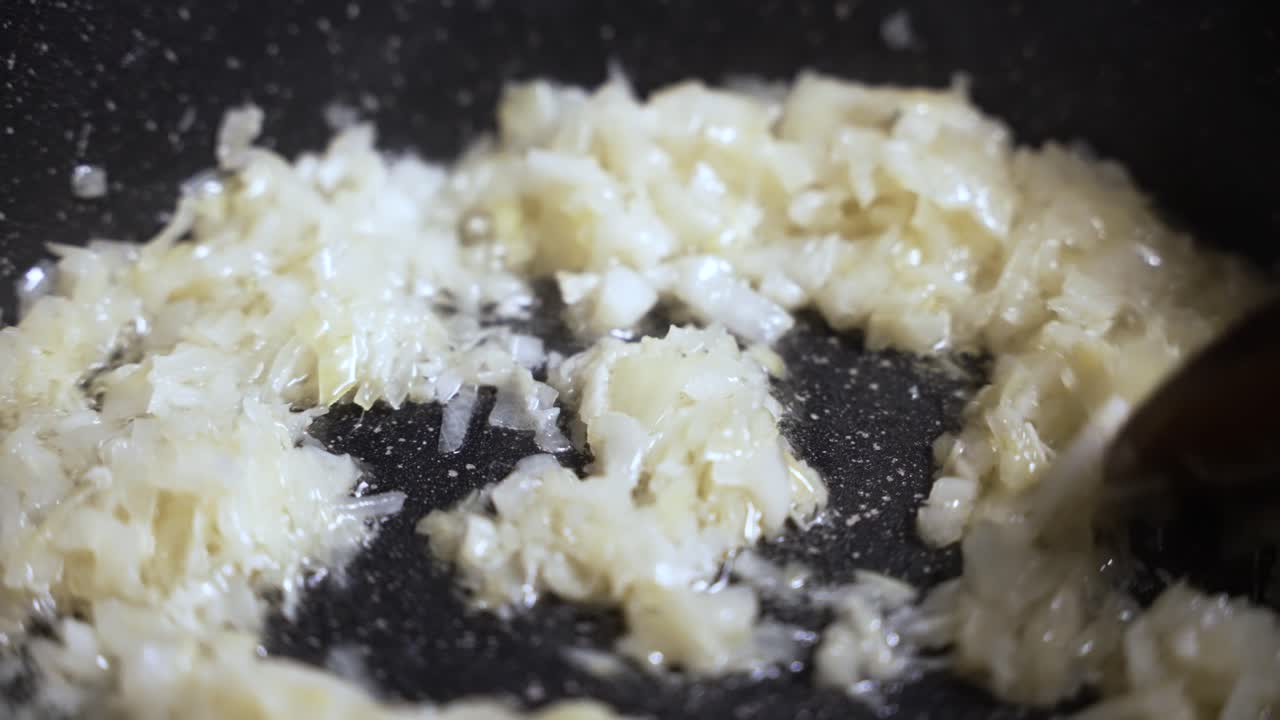 Close up of chopped white onions frying in hot oil in a black pan, creating steam and sizzling sounds