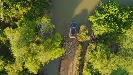 aerial top down view on black suv off-road vehicle on a green forest road passing through deep mud puddle on sunny day in africa