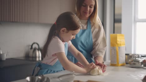 curious little girl is helping mother to cook in kitchen, learning to knead dough on table, cooking pie