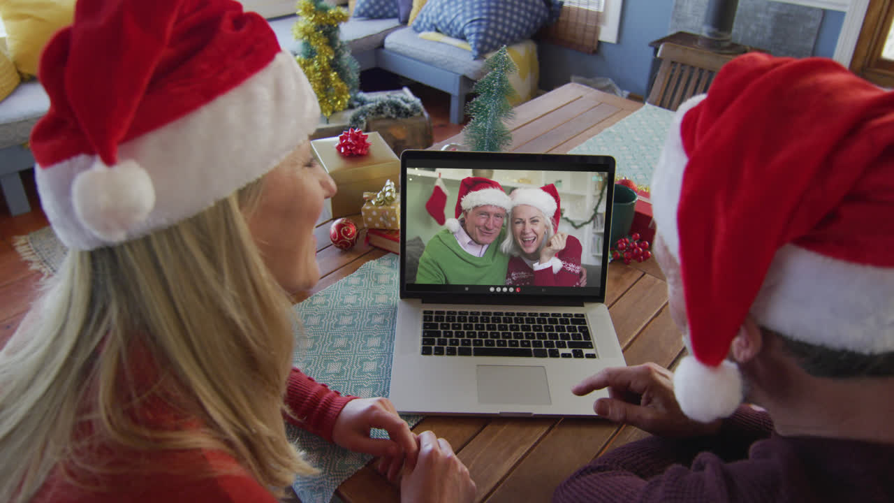 pareja caucásica con sombreros de santa usando una computadora portátil para una videollamada de navidad con la pareja en la pantalla
