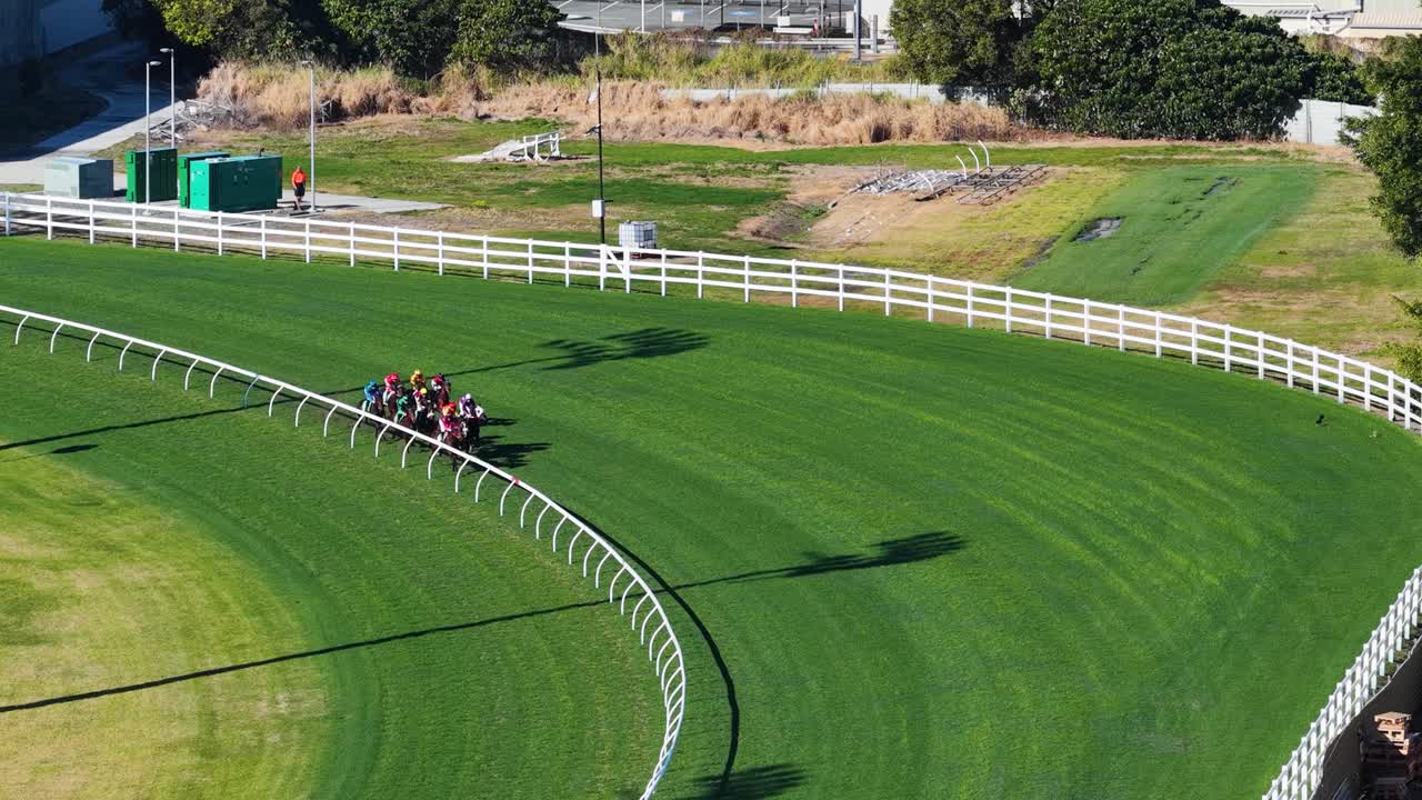 Multiple jockeys ride racehorses around a green turf track curve in bright daylight, captured from above with smooth drone camera movement