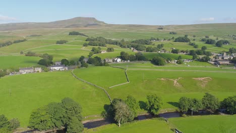 drone footage, moving and panning showing the mountain of ingleborough in the distance with a rural yorkshire countryside village and river on a sunny summer day