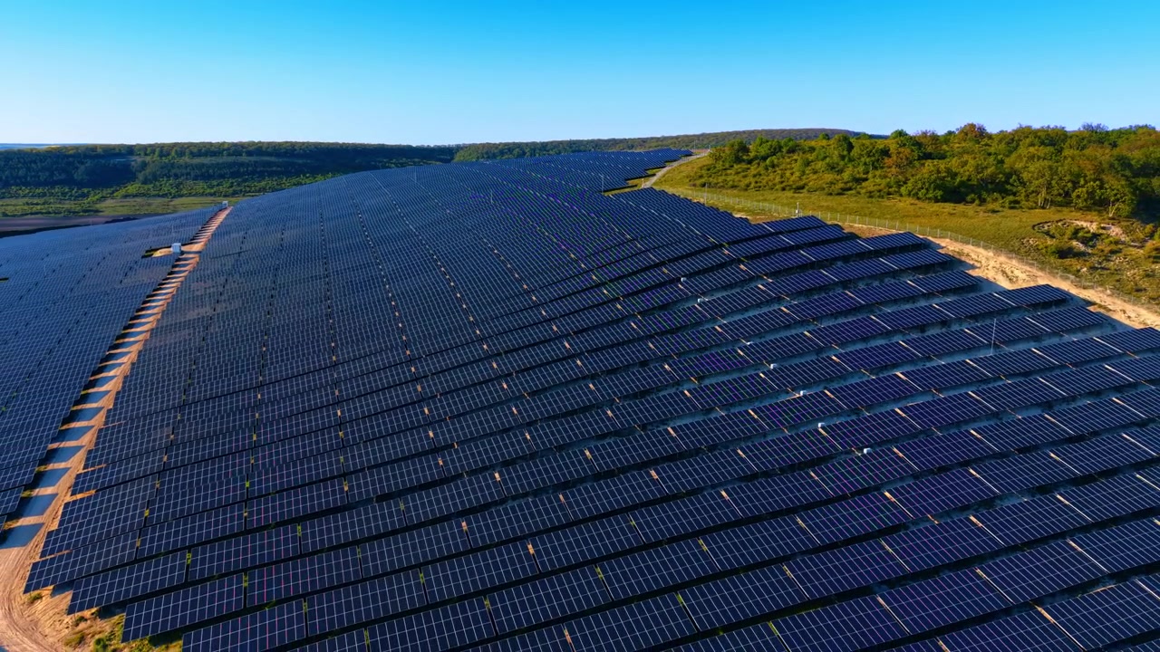 Tæt solcelleanlæg dækker en bakketop med skovkant. Diagonal luftfoto af mørke panelrækker på bølgende terræn med grøn trækant og blå himmel
