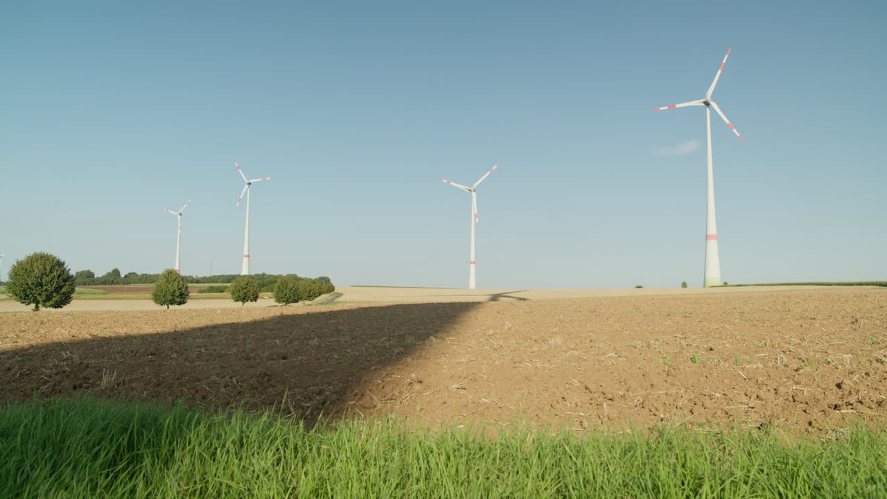 Four large wind turbines stand on open farmland with fresh green grass in the foreground, casting long shadows under a bright blue sky