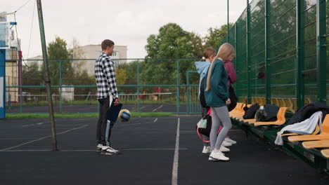 coach holding performance book and bouncing volleyball while waiting as students place bags on bench in volleyball court, trees and buildings in background