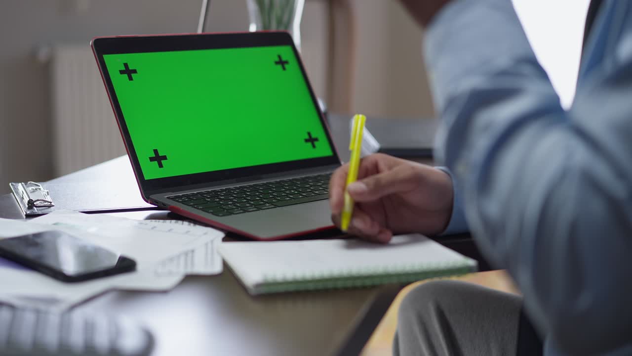 Laptop with chromakey green screen on table with male African American hands writing filling in documents. Unrecognizable young businessman working online in home office and drinking tea.
