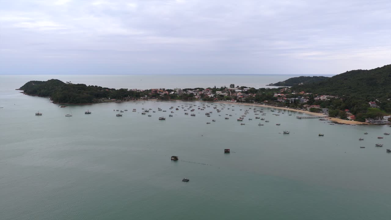 High-altitude drone shot approaches Praia do Trapiche, showcasing numerous fishing boats docked in the water amid cloudy afternoon