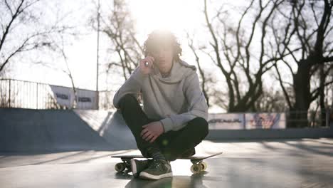 Der-Junge-Mann-Ruft-Freunde-Mit-Dem-Handy-An,-Während-Er-In-Einer-Skatezone-Im-Skatepark-Sitzt