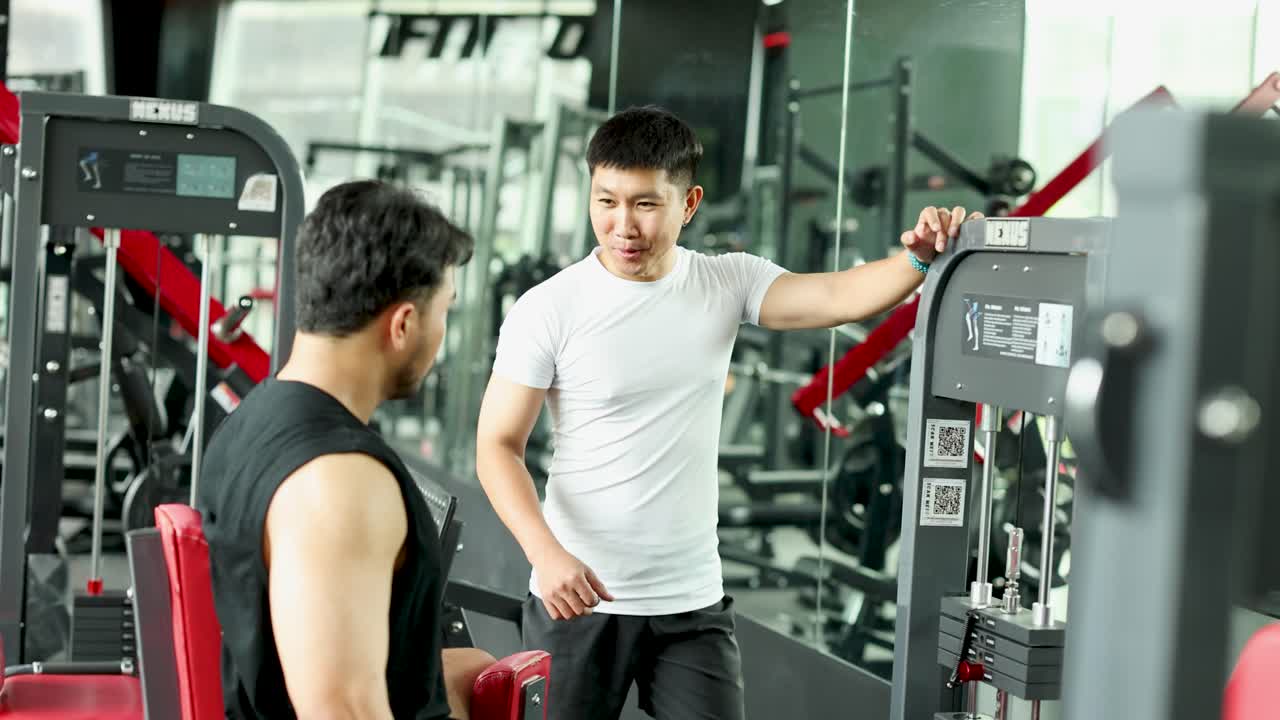 Two men engage in a lively conversation in a gym setting, surrounded by exercise equipment and bright lighting