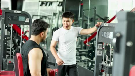 two men engage in a lively conversation in a gym setting, surrounded by exercise equipment and bright lighting