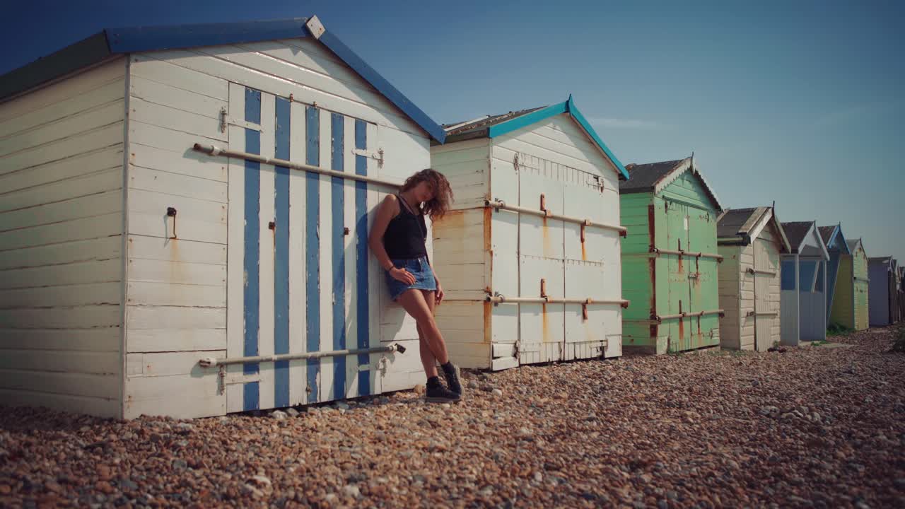bucle de video sin inconvenientes: cinemagrafía de una joven modelo morena con falda corta apoyada en coloridas cabañas de playa en brighton, al sur de inglaterra, con el cabello moviéndose suavemente con el viento