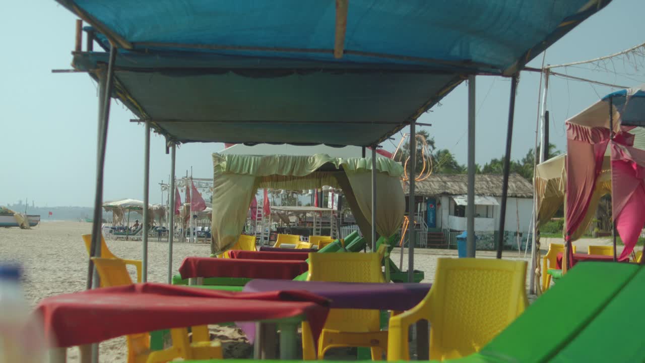 Sedie e tavoli colorati vuoti disposti sotto baldacchini ombreggiati su una spiaggia sabbiosa, creando un'atmosfera rilassata in riva al mare durante una giornata tranquilla.