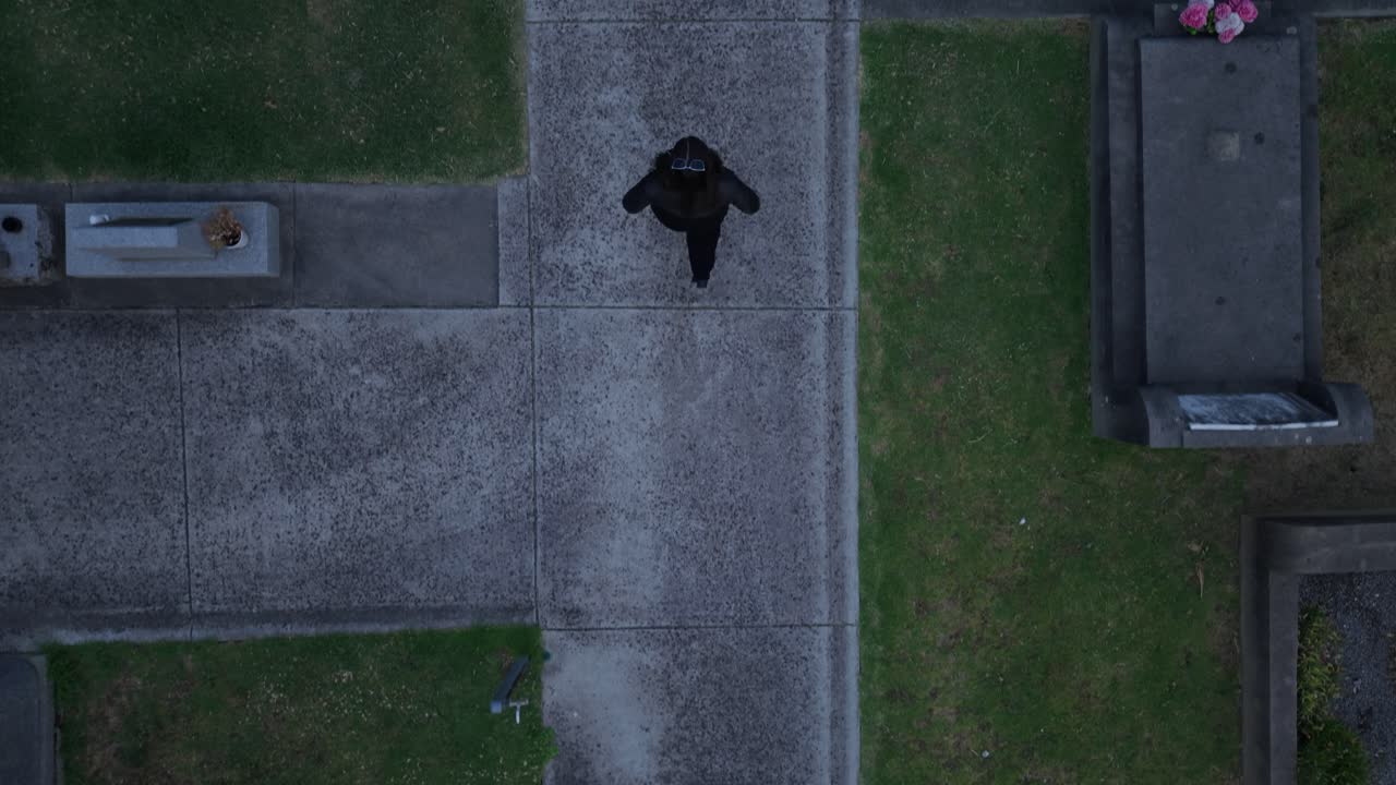 Riprese drone che mostrano una ragazza solitaria camminare attraverso un cimitero al tramonto a Ferntree Gully, Melbourne. La scena è tranquilla, con lunghe ombre e un'atmosfera cupa.