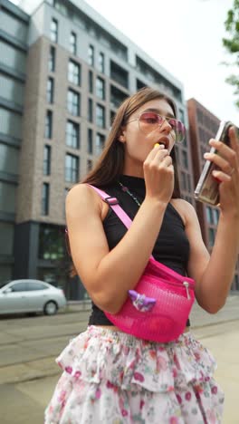 teenage girl applying lipstick while looking at smartphone on city street