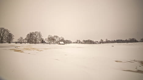 Tiro-De-Lapso-De-Tiempo-De-La-Nieve-Que-Cubre-El-Suelo-Que-Se-Derrite-Lentamente-A-Lo-Largo-Del-Campo-Rural-Con-La-Vista-De-La-Cabaña-Del-Pueblo-En-El-Fondo