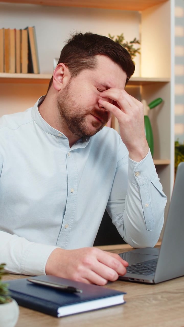 Giovane uomo al tavolo di casa con mal di testa che si massaggia le tempie mentre lavora al laptop dopo un burnout, al chiuso.