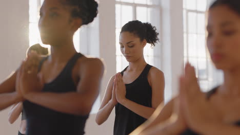 young mixed race yoga woman practicing prayer pose meditation with group of multiracial women enjoying healthy lifestyle exercising in fitness studio at sunrise