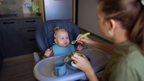 mother sits at the high chair with her baby. woman takes some avocado on the spoon and gives it to child.
