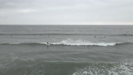 Surfer-In-Neoprenanzügen-Fangen-An-Einem-Flachen,-Bewölkten-Ozeantag-Eine-Kleine-Graue-Welle