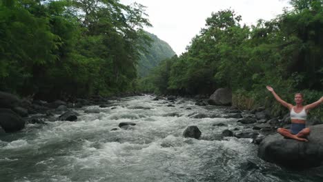 one with nature concept, woman meditating in lotus pose on rock, river flowing