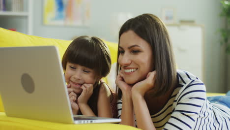 Glückliche-Mutter-Und-Tochter,-Die-Sich-Etwas-Auf-Dem-Laptop-Ansehen,-Der-Auf-Dem-Gelben-Sofa-Im-Wohnzimmer-Liegt