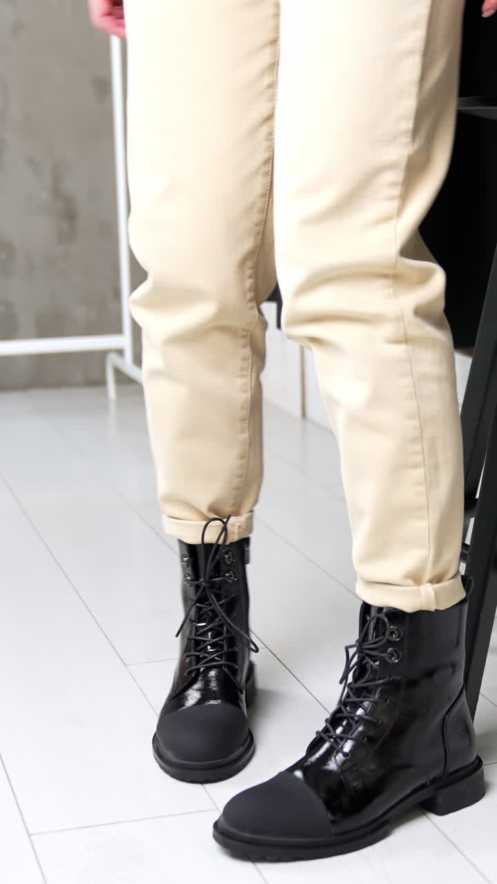 Unknown woman comes up to a high stool and sits down. Model demonstrates stylish boots from combined leather. White backdrop. Vertical video