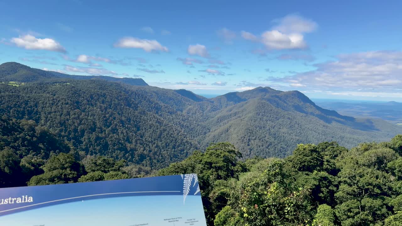 Camera pans across a scenic lookout sign and lush rainforest mountains under bright daylight, revealing expansive views of Dorrigo National Park, New South Wales, Australia