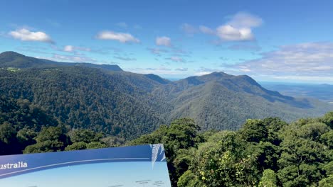 camera pans across a scenic lookout sign and lush rainforest mountains under bright daylight, revealing expansive views of dorrigo national park, new south wales, australia