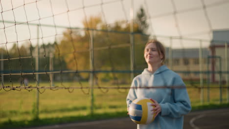 joven con capucha jugando al voleibol en la cancha al aire libre, centrada en la acción, sosteniendo el voleibol, con la red visible en primer plano y la vegetación borrosa y el edificio en el fondo
