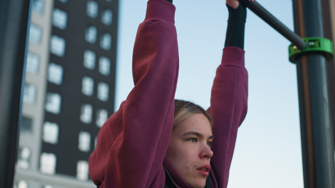 Workout participant grips pull up bars lifting legs with focus and effort, engaging in intense exercise outdoors, children on rollers seen playing on the other side of the park