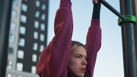workout participant grips pull up bars lifting legs with focus and effort, engaging in intense exercise outdoors, children on rollers seen playing on the other side of the park