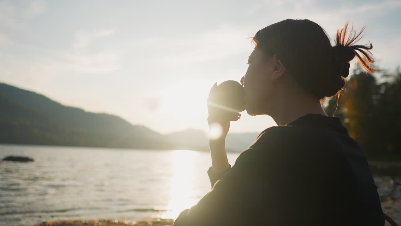 해가 지는 동안 호수 근처에서 사과를 먹는 여자, 외로운 여자가 산과 호수의 전망을 즐기며 과일을 먹는 여성, 은 가을 날, 주말 휴식