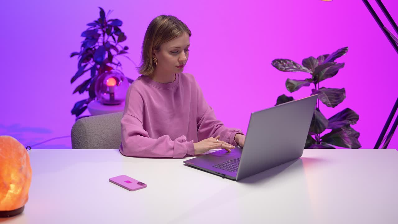 Woman in a pink sweatshirt working intently on a laptop at a white desk in a vibrant home office filled with soft neon lighting, stylish plants, and modern decorative elements creating a cozy ambiance