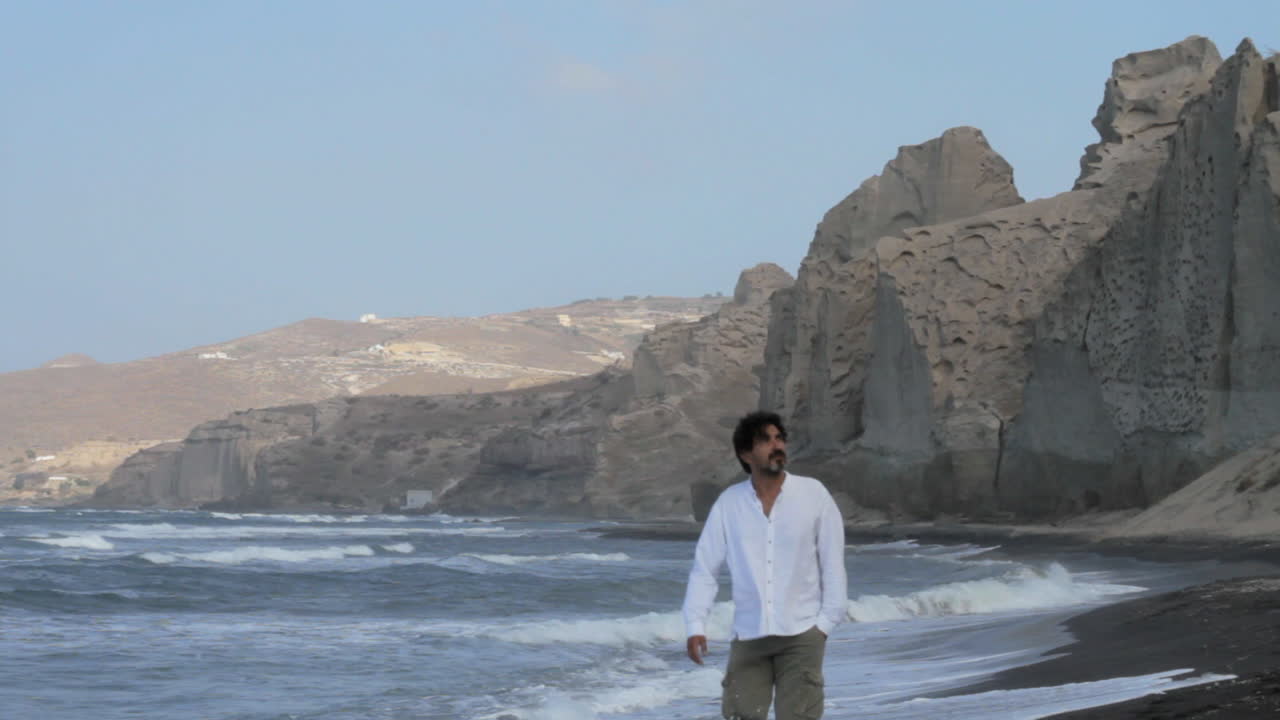 un hombre camina solo en una playa de arena negra rodeada de formaciones de acantilados volcánicos blancos en la isla de santorini, grecia