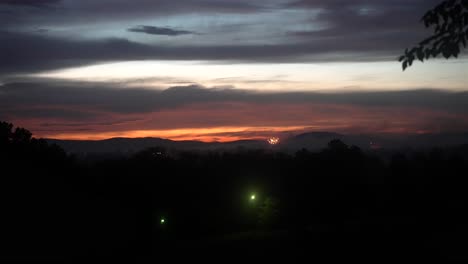 wide angle overview of fireworks launching into sky above forested mountains at dusk in slow motion